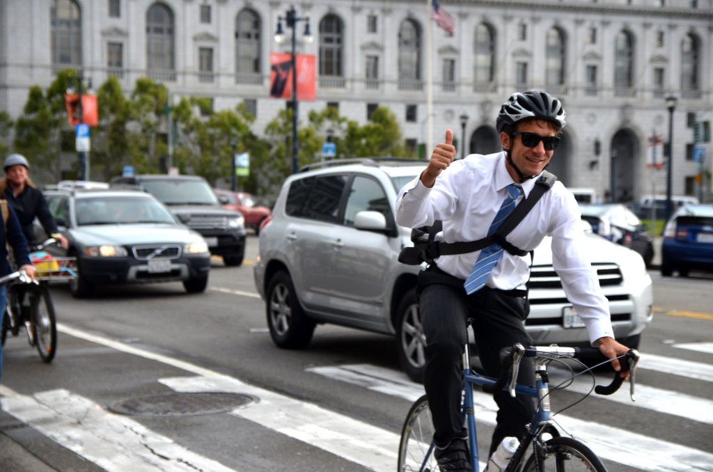 A bike commuter wearing a suit, tie, and a helmet flashes a thumbs up to the photographer while biking on a busy road in San Francisco. 