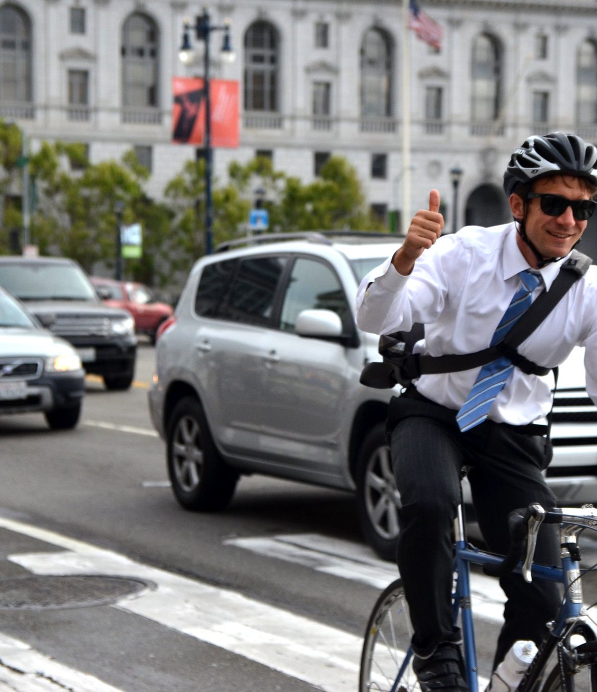 A bike commuter wearing a suit, tie, and a helmet flashes a thumbs up to the photographer while biking on a busy road in San Francisco.