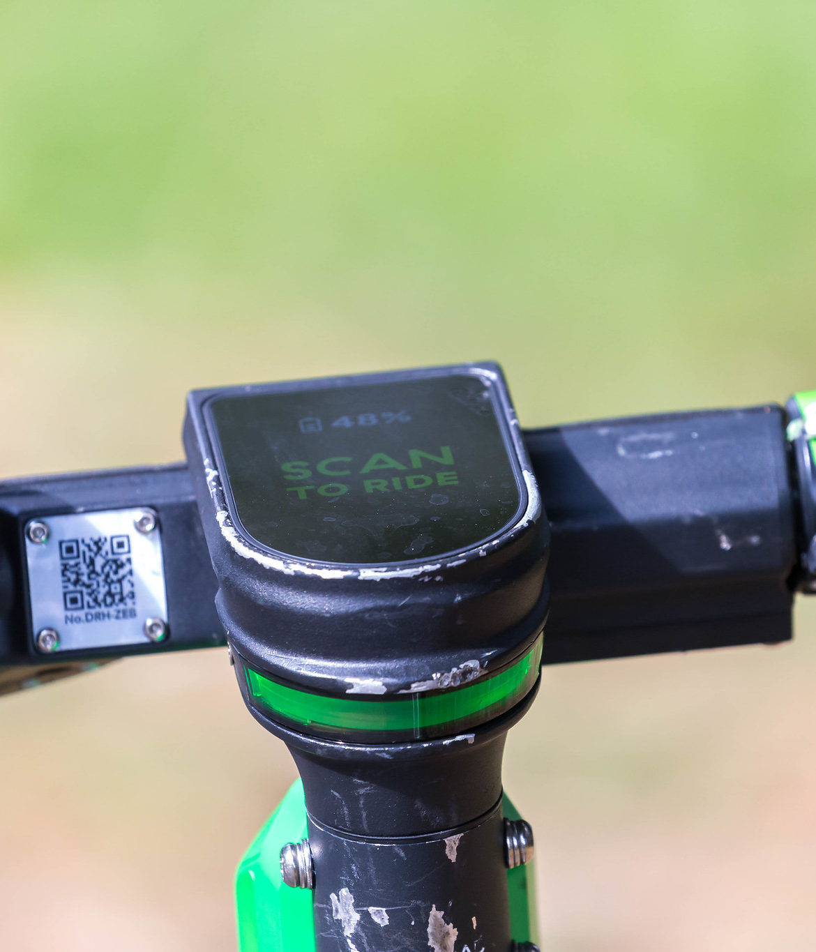 A close-up of the handlebars of a Lime electric scooter. The scooter has a small computer screen that reads "Scan to Ride". To the right of the screen is a QR code.