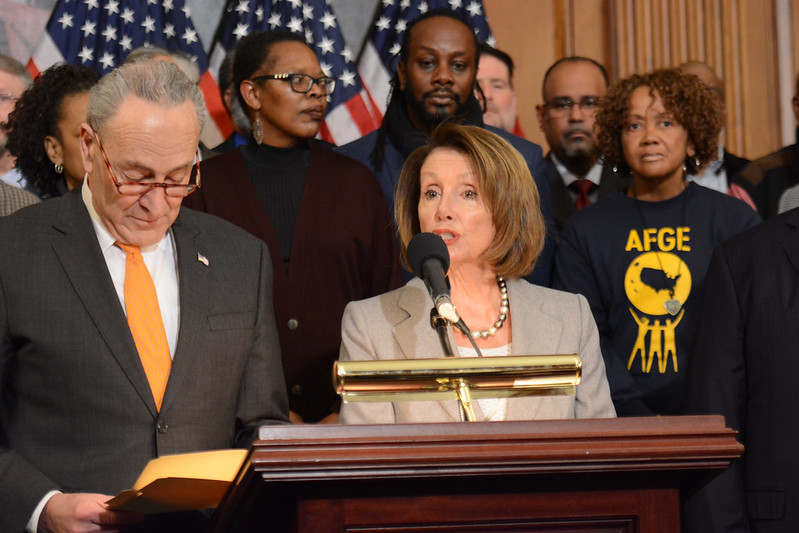 Nancy Pelosi speaking into a microphone with Chuck Schumer on her right, AFGE behind her