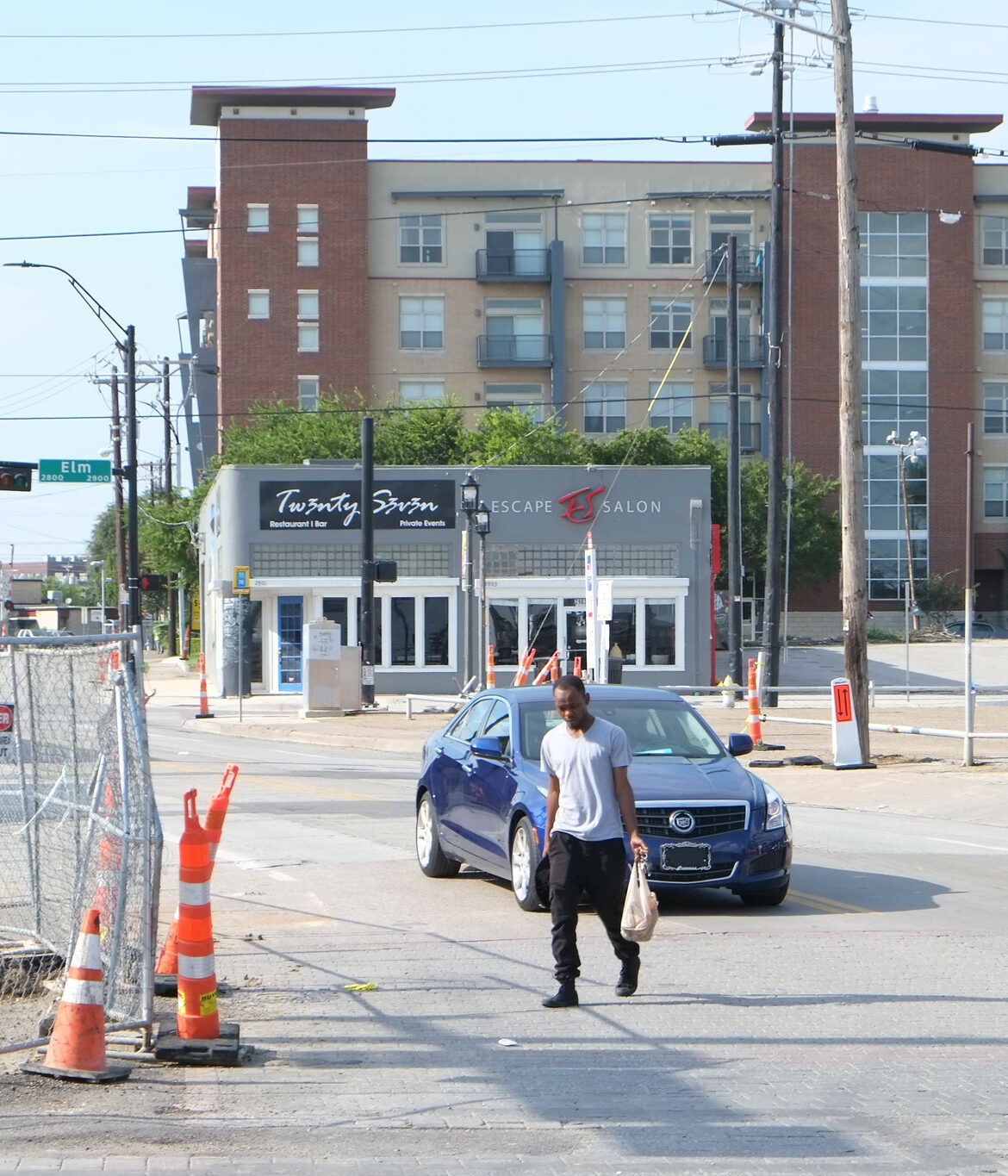 A Black man crosses a street without a crosswalk carrying grocery bags