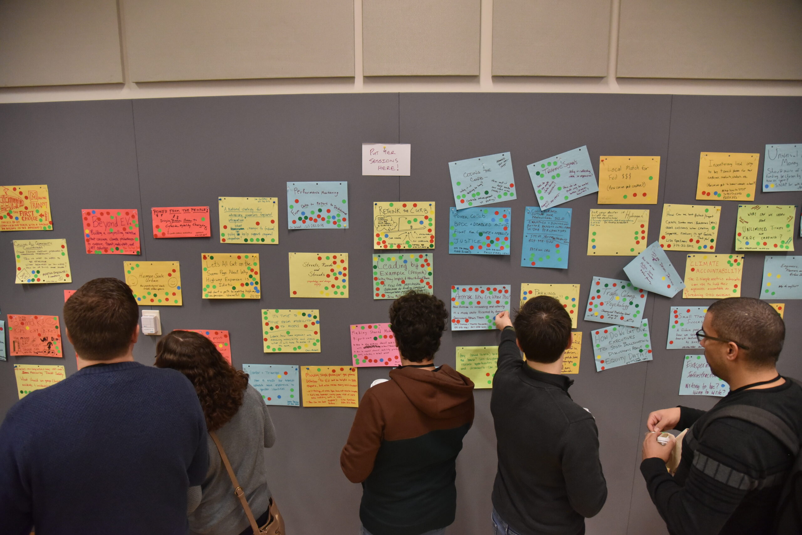 backs of people at tcamp sticking sheets of paper with session proposals on the board