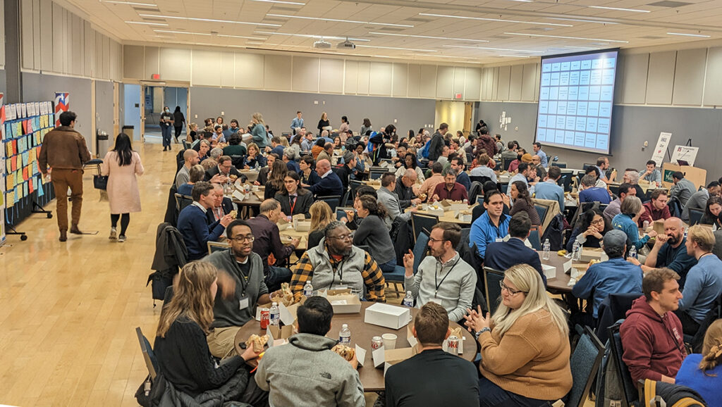 a wide shot of a full room of people around round tables eating lunch and talking