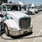 A line of electric trucks wait to be charged in a wide, half-empty parking lot