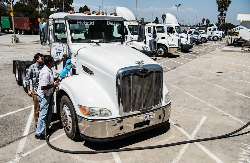 A line of electric trucks wait to be charged in a wide, half-empty parking lot