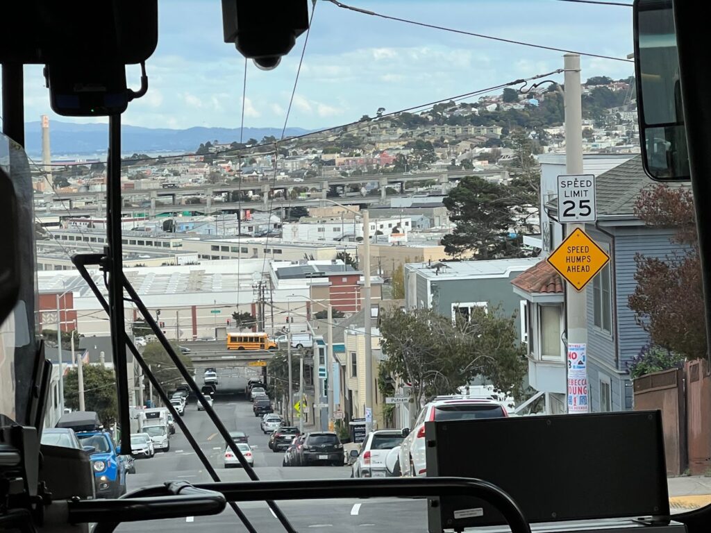 Picture taken through a bus front window of a street lined with vehicles leading to the hills of Bayview and Hunters Point