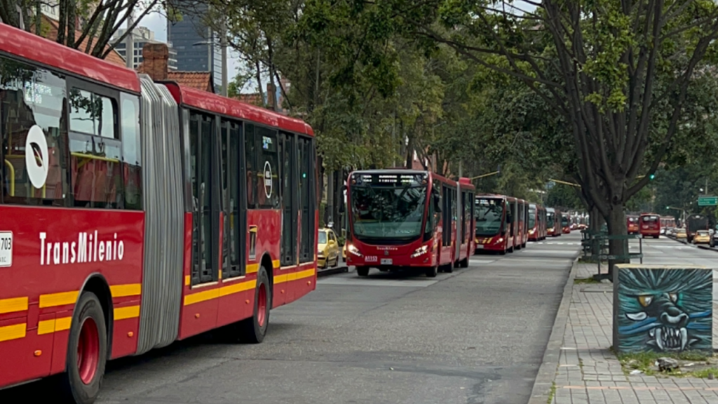 A line of six red TransMilenio buses stream down a tree-lined street