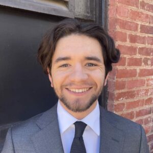 Corrigan smiles approachably in a professional suit and tie in front of a black and brick wall.