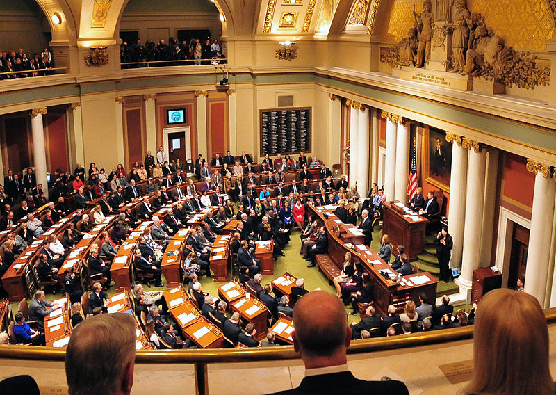 Members of the Minnesota legislature assemble in a warmly lit space with gold decorations and white columns