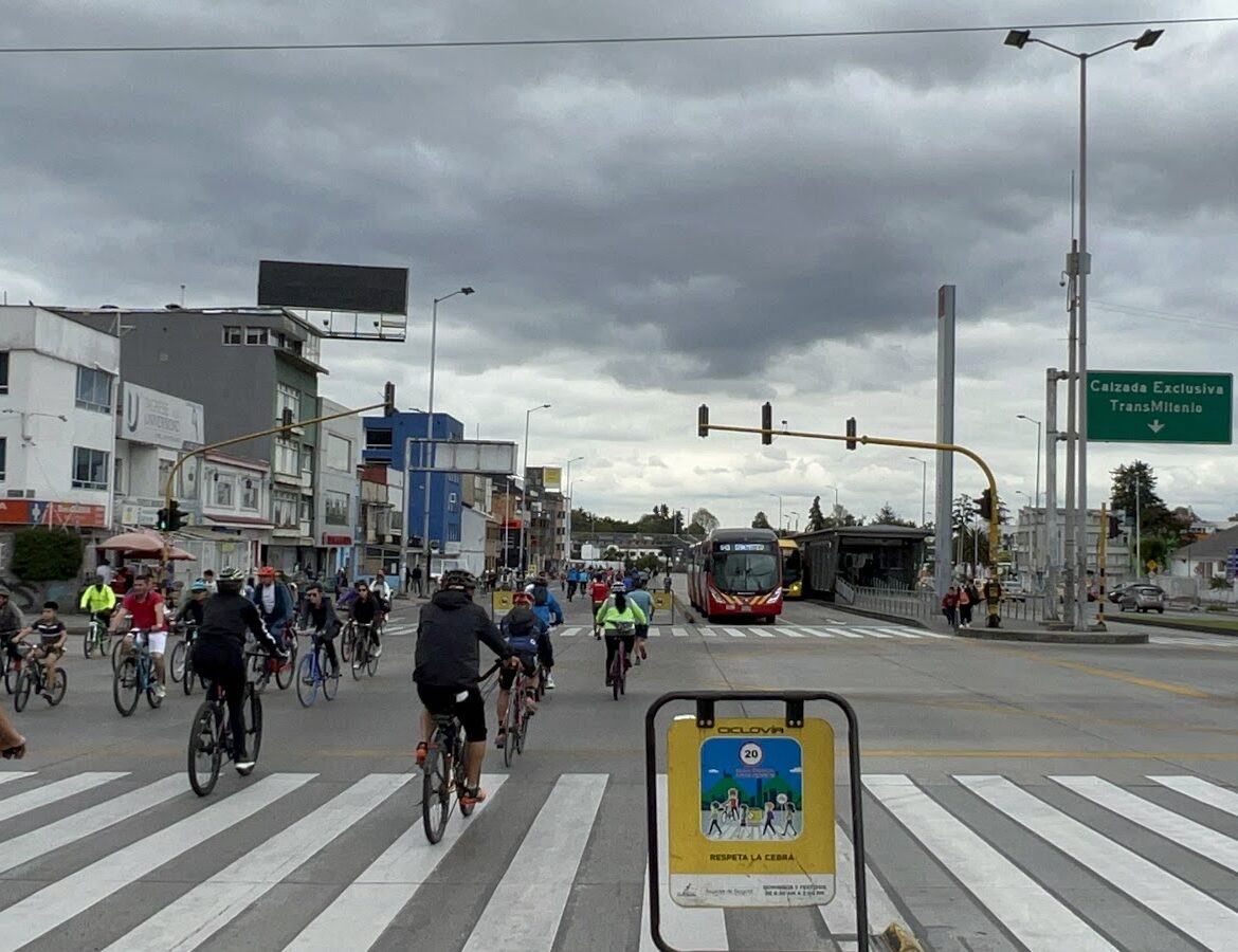A full range of transportation options are on display: bicyclists and pedestrians in the left lane, a bus in the right lane, and wide crosswalks in the foreground.