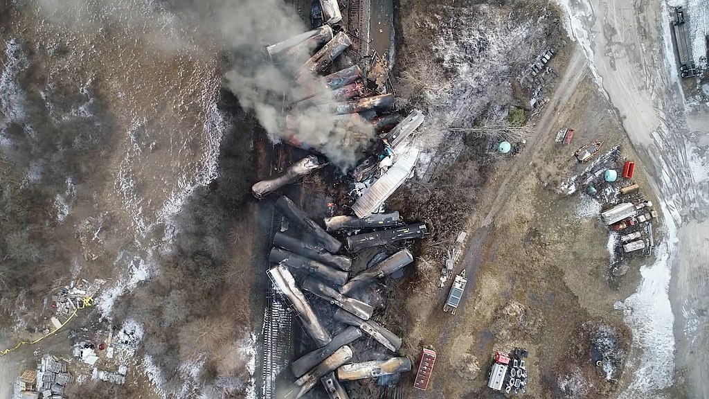 Aerial view of several rolled train cars piled on top of and near each other, with smoke, debris, and water surrounding them.
