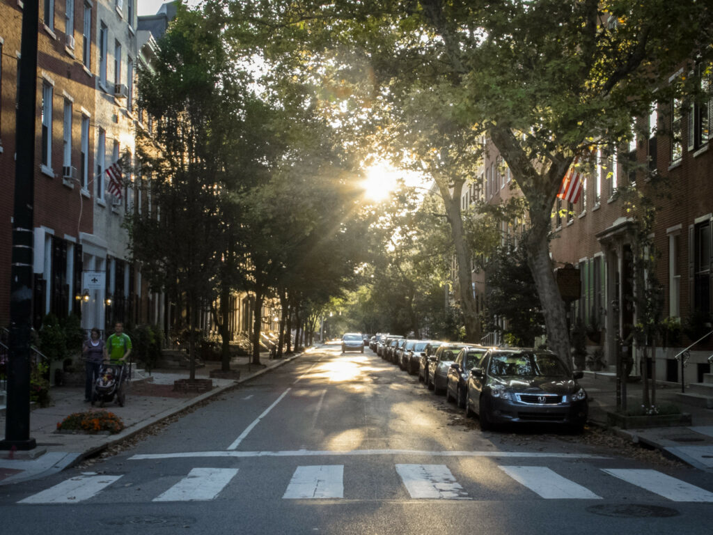 A man rolls a stroller down a wide sidewalk along a tree-lined street with a painted bike lane and crosswalk