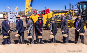 a line of people in suits digging from a pile of dirt for a ceremonial groundbreaking for a new highway