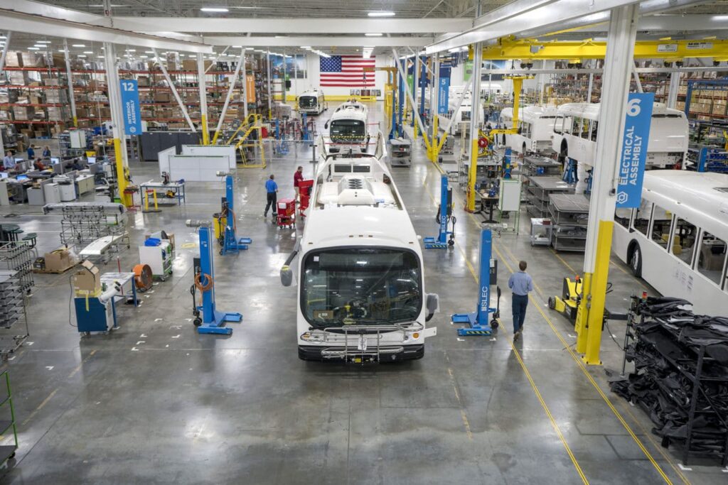 Electric buses line up in a  brightly lit warehouse with an American flag in the background