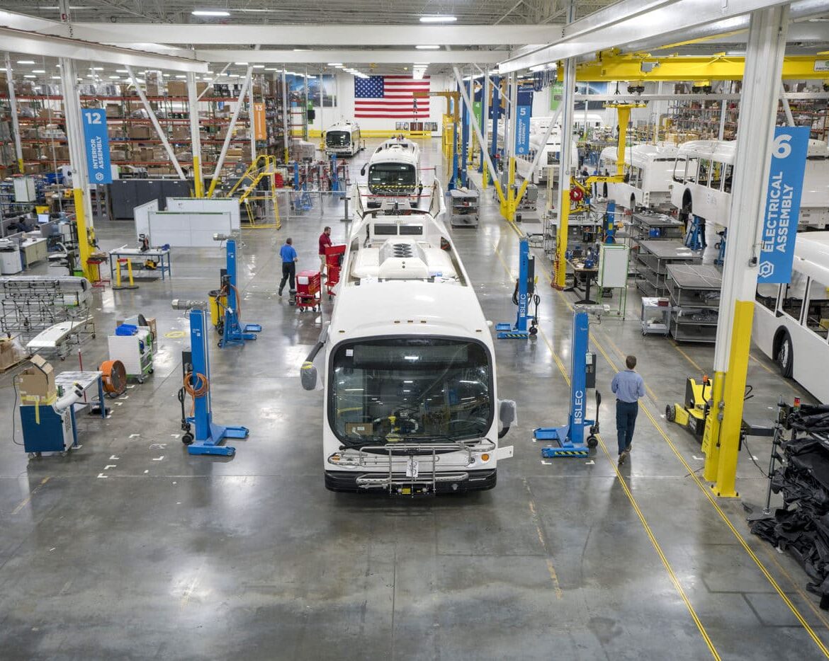 Electric buses line up in a brightly lit warehouse with an American flag in the background