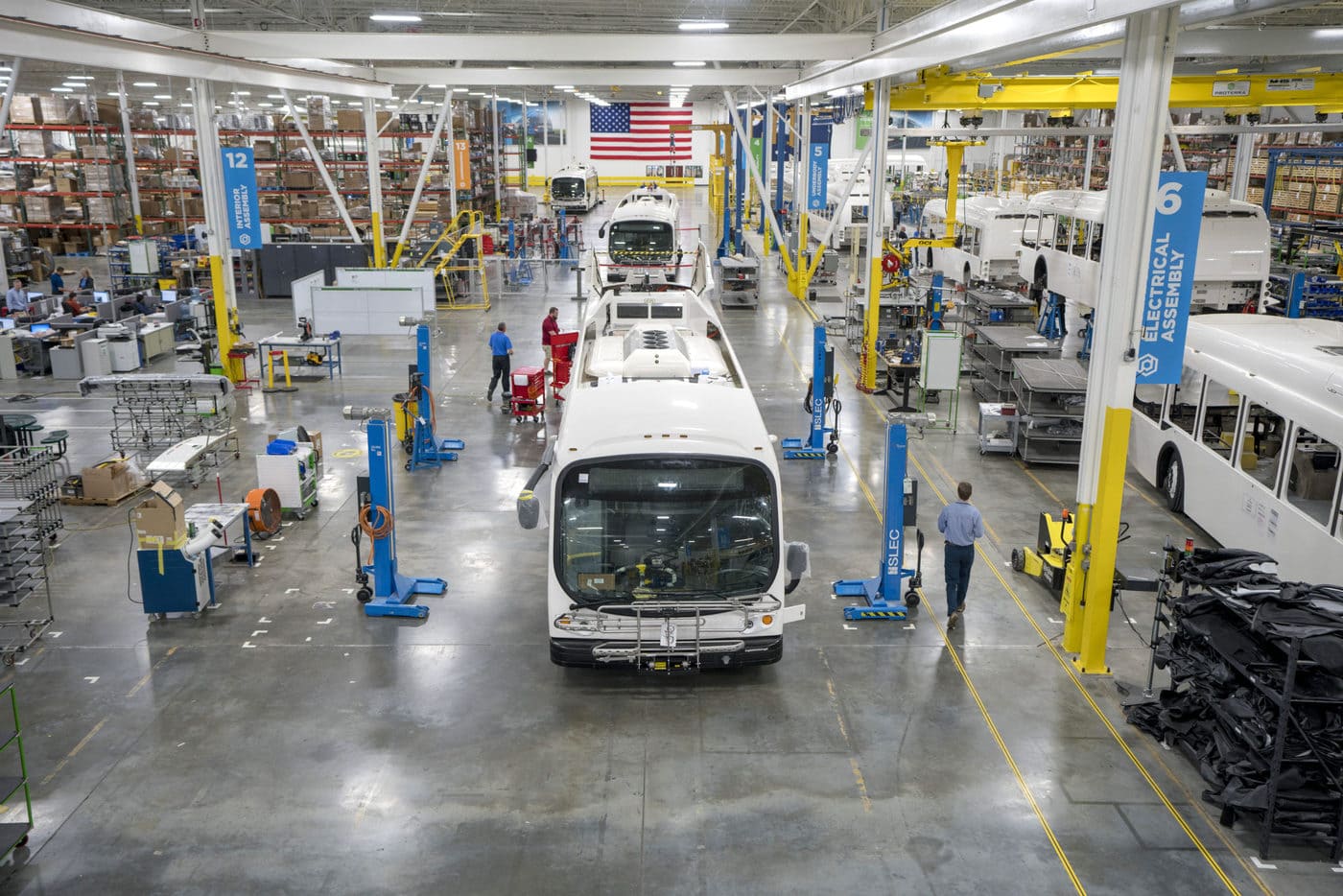 Electric buses line up in a brightly lit warehouse with an American flag in the background