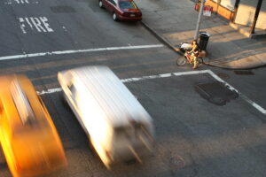 A young woman holds onto her bicycle, waiting for the ped signal to cross a crosswalk showing signs of wear.