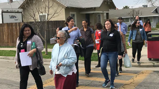 A group of people representing a range of ages, genders, and ethnicities walk across a cracked road within a marked crosswalk.