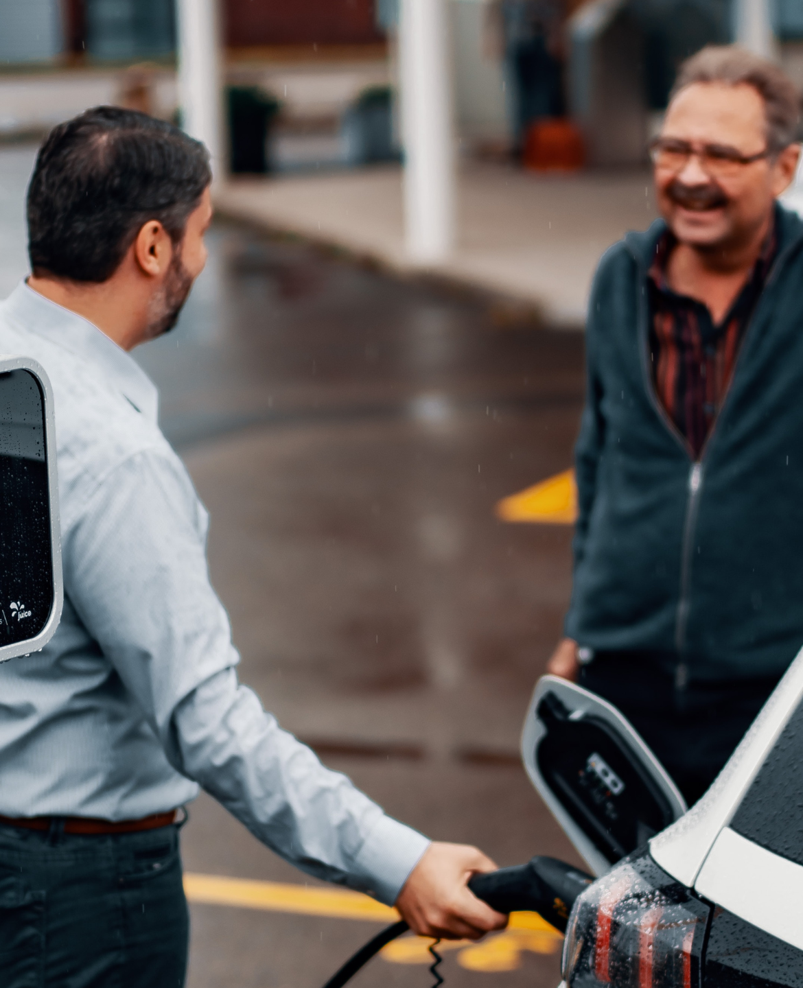 Two men stand, chatting, beside a car while it's getting plugged in to charge.