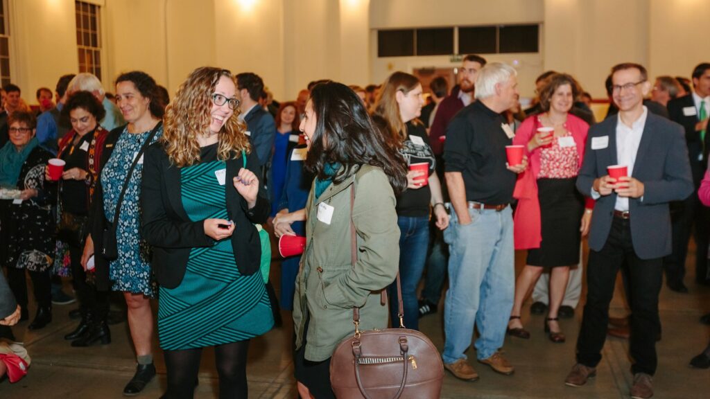 A group of people in business professional attire gathers in a room talking and smiling over drinks