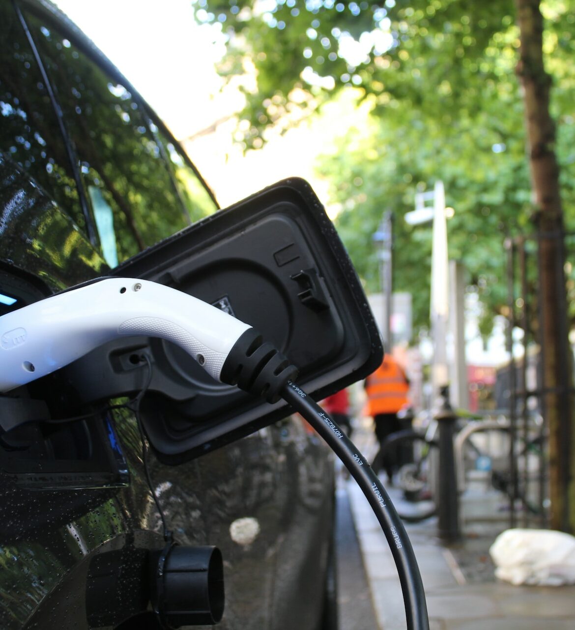 A black EV charges on the side of a tree-lined street. In the background, a construction crew in orange vests mingles on a wide sidewalk with bike parking.