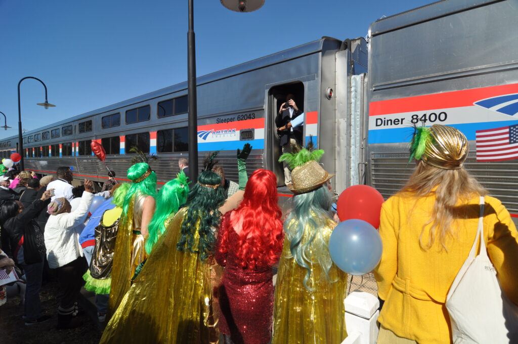 the backs of women in colorful wigs and costumes looking at amtrak train in background