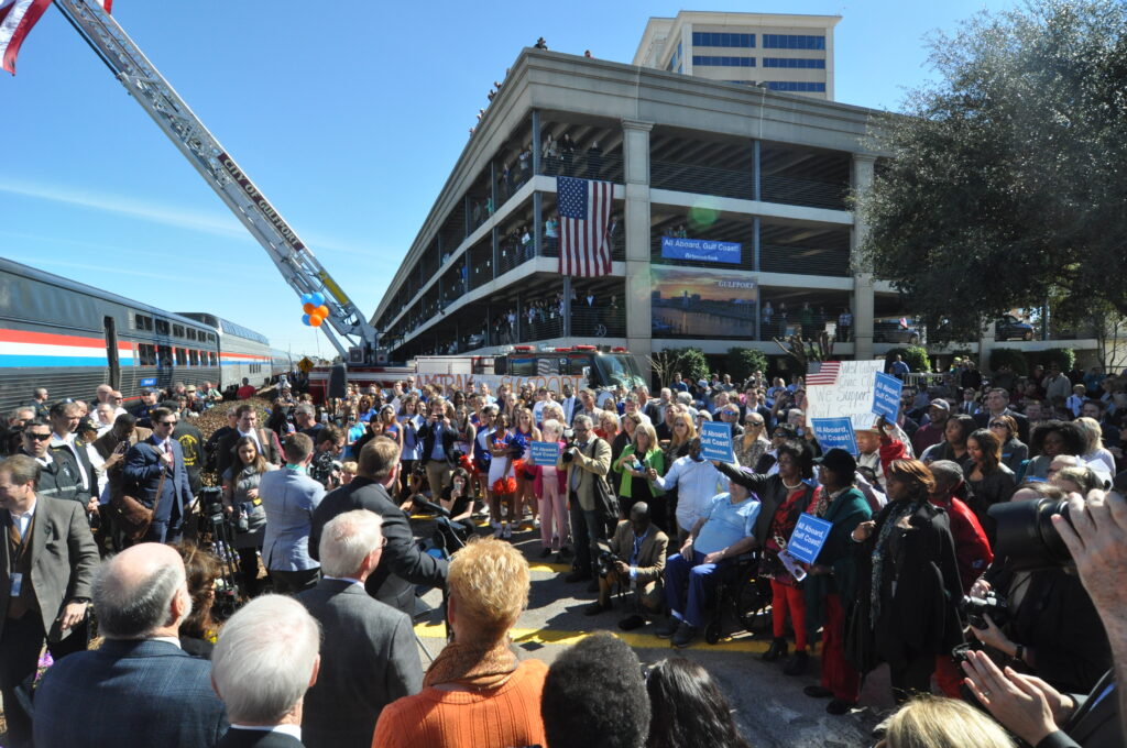 wide shot showing big crowd of people with parking garage behind and amtrak train at left