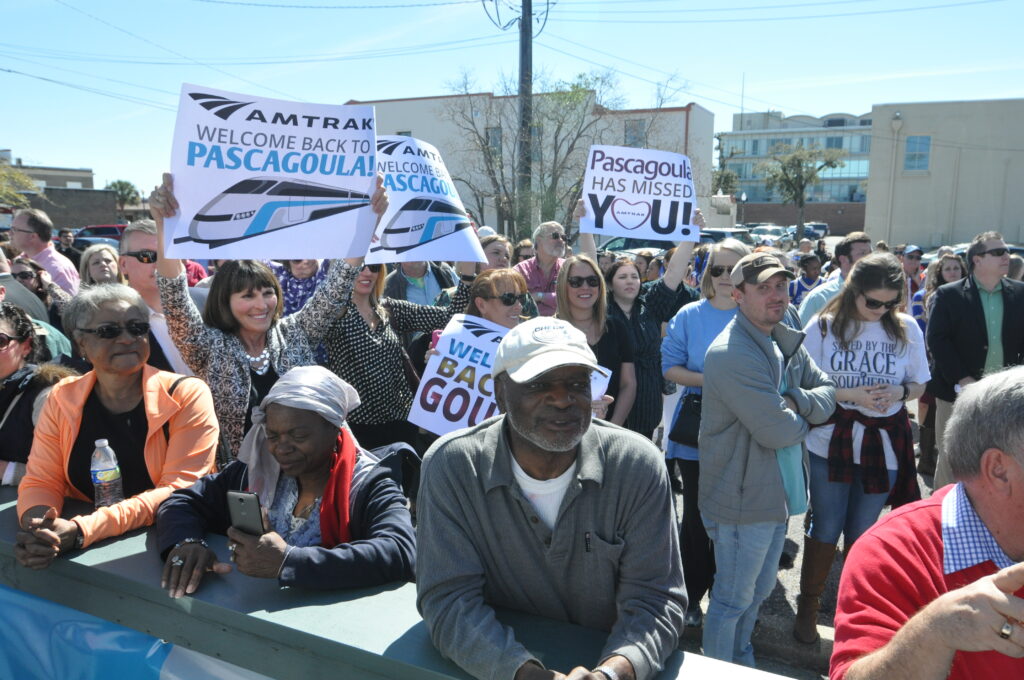 closeups of people, some with signs reading "Amtrak - welcome back to Pascagoula"