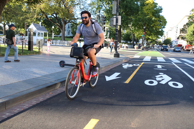 A man with dark hair and sunglasses rides a Capitol Bikeshare bicycle down a painted bike lane near a treelined sidewalk.