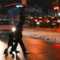 An adult and small child cross the street at night without a crosswalk while cars approach