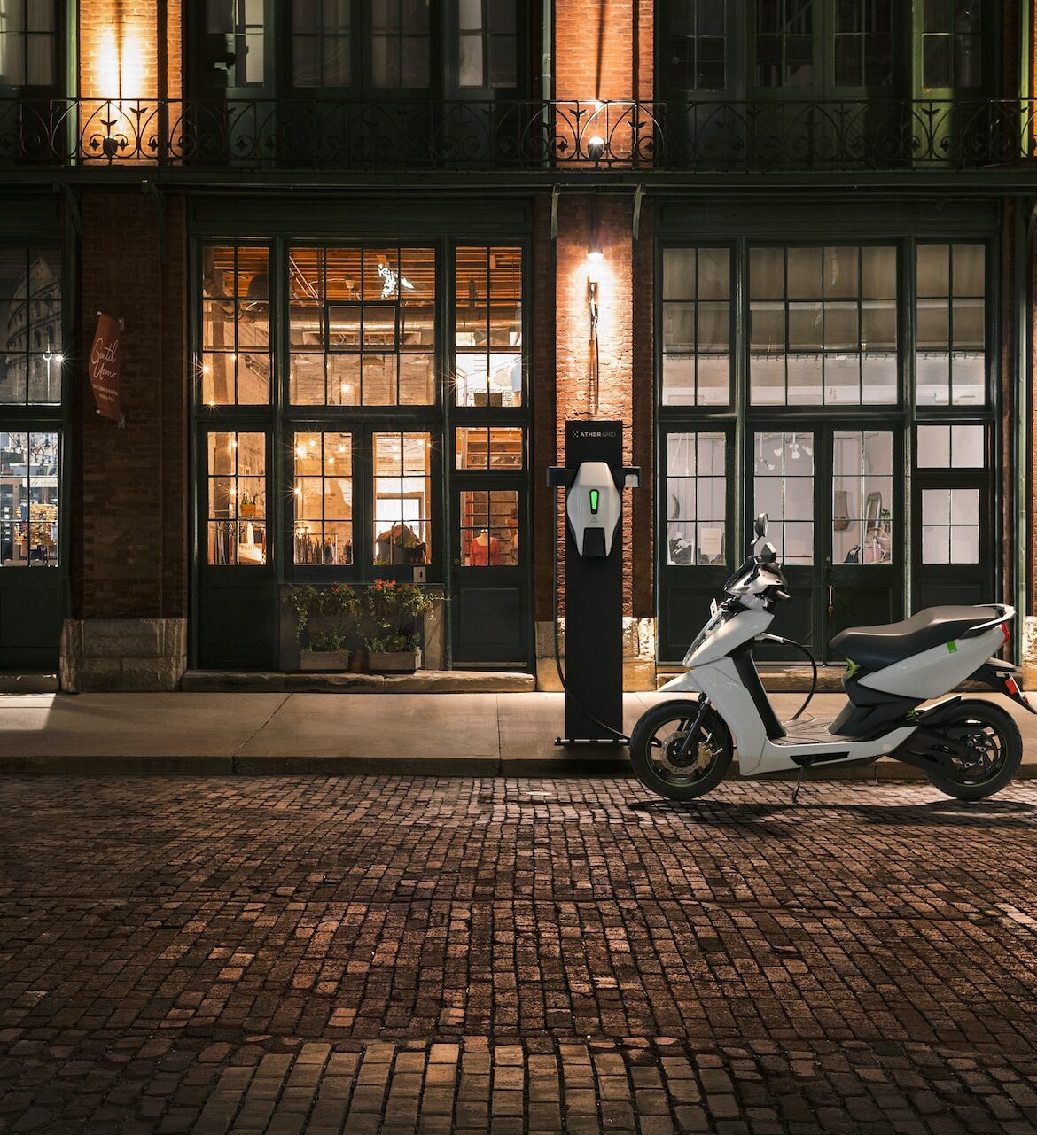 An electric scooter charges at the curb in front of a warmly lit storefront at night