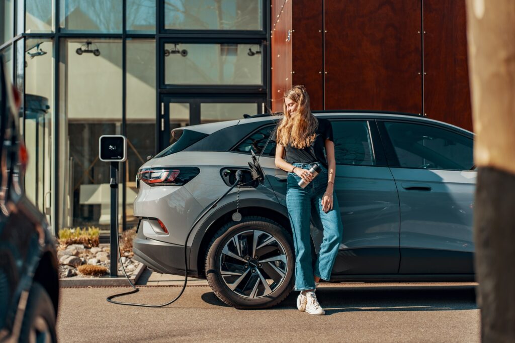 A woman leans against her EV while it charges outside of an apartment building