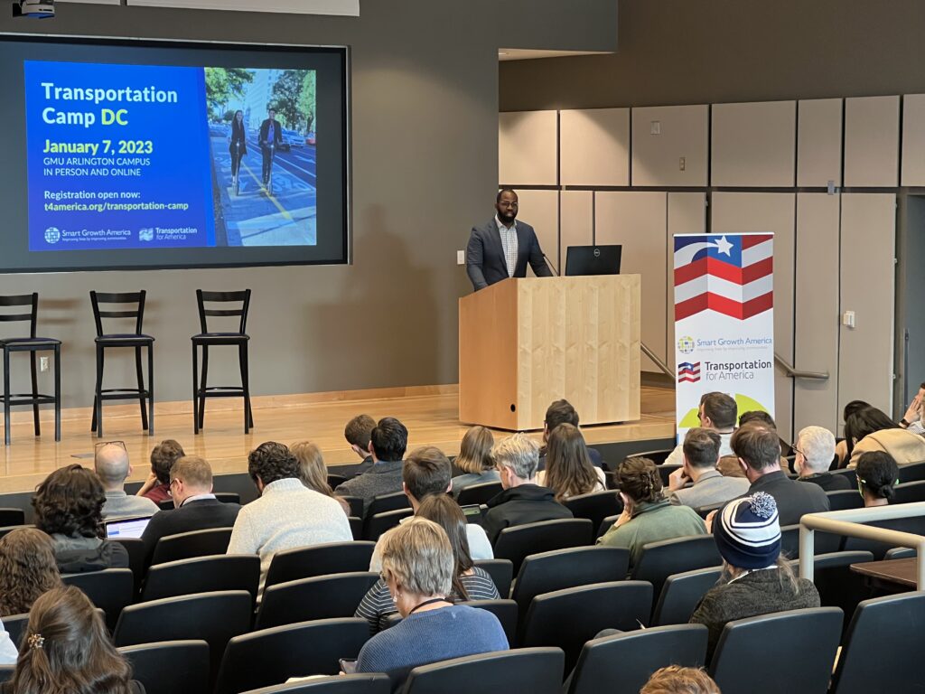 A Black man in a suit stands behind a podium. The slide behind him reads "TransportationCamp DC January 7, 2023, GMU Arlington Campus in person and online"