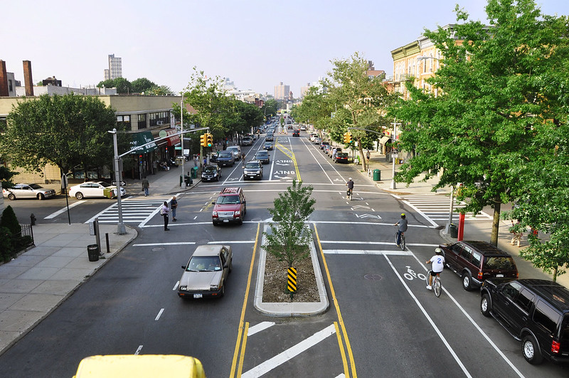 A calm tree-lined street in Brooklyn, NY hosts one lane of car traffic, a bike lane, street parking, and a median to shorten the crosswalk distance for pedestrians.