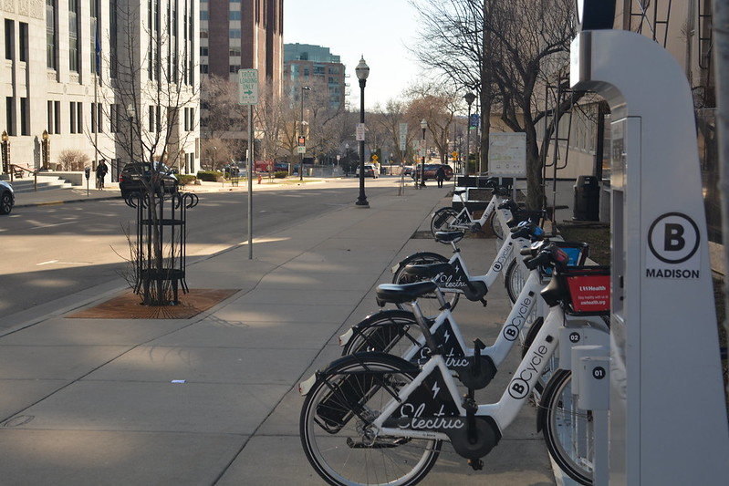 electric bikes line up at a docking station on a wide sidewalk in madison, wisconsin
