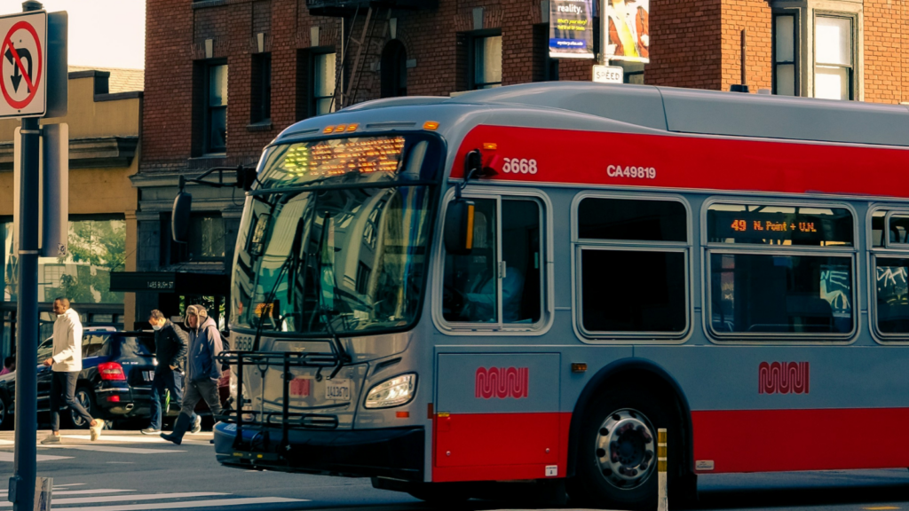 A bus rolls to a stop on a bustling city street as pedestrians walk down a wide sidewalk in the background