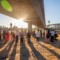 A group of people representing a range of ages, genders, and ethnicities, stands in a circle beneath a highway overpass, with the sun rising in the background