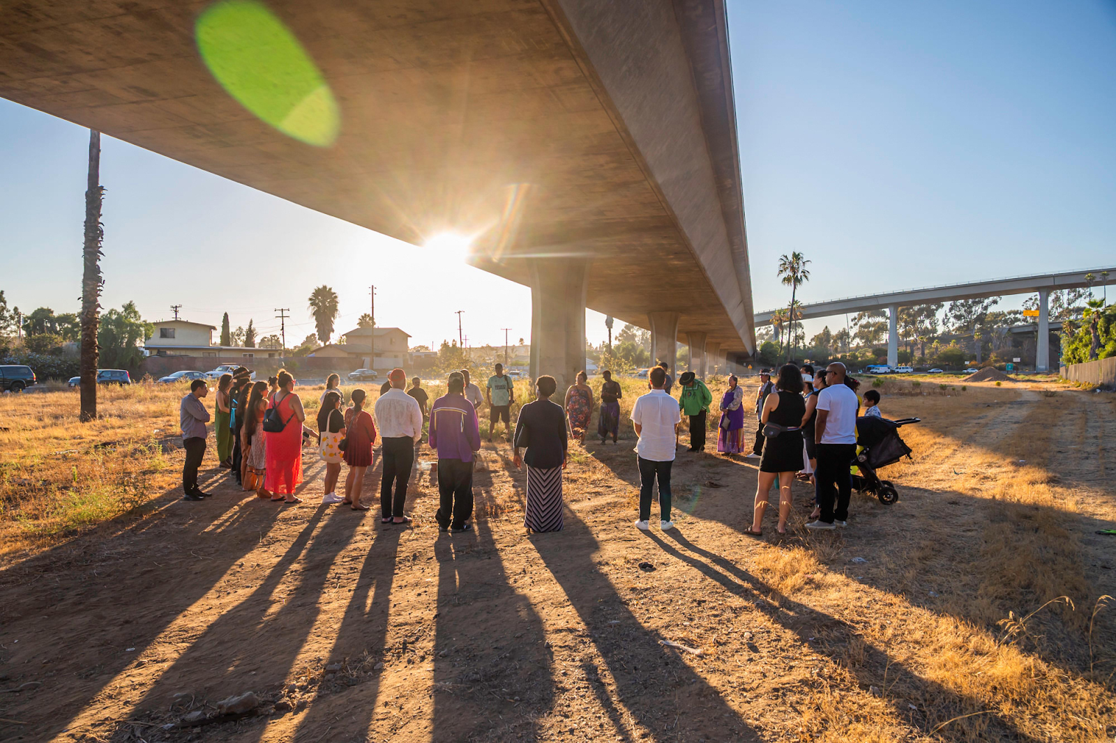 A group of people representing a range of ages, genders, and ethnicities, stands in a circle beneath a highway overpass, with the sun rising in the background