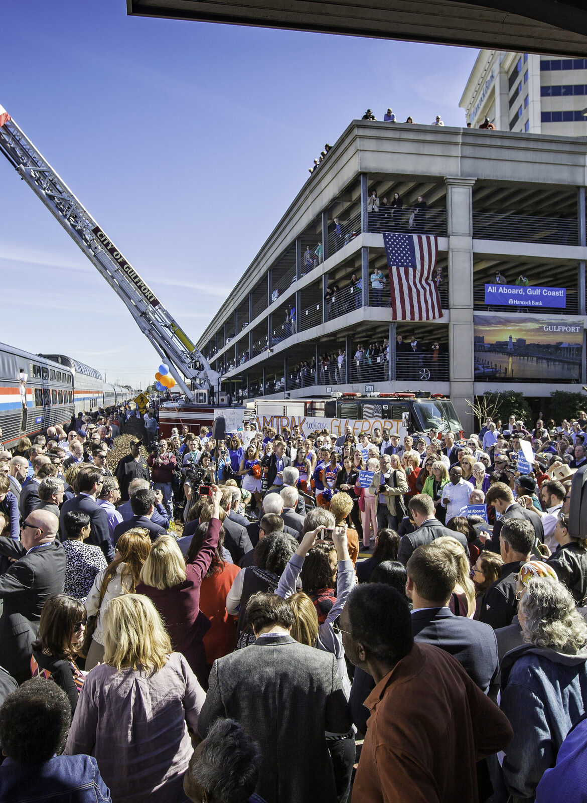 A crowd of people gathers by an Amtrak train, a U.S. flag waving above them.