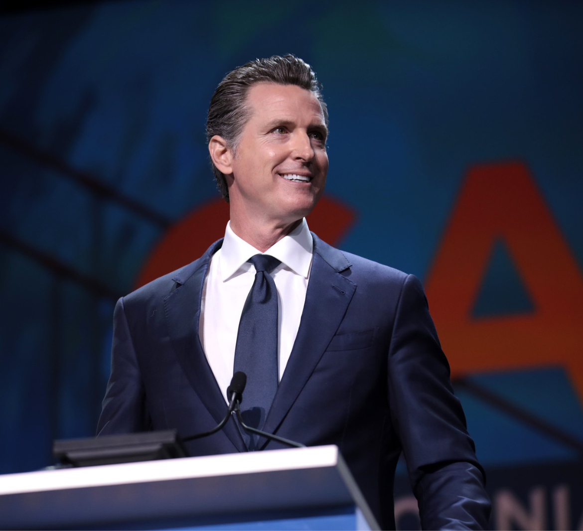 Governor Gavin Newsom wears a blue suit and tie, smiling from a podium