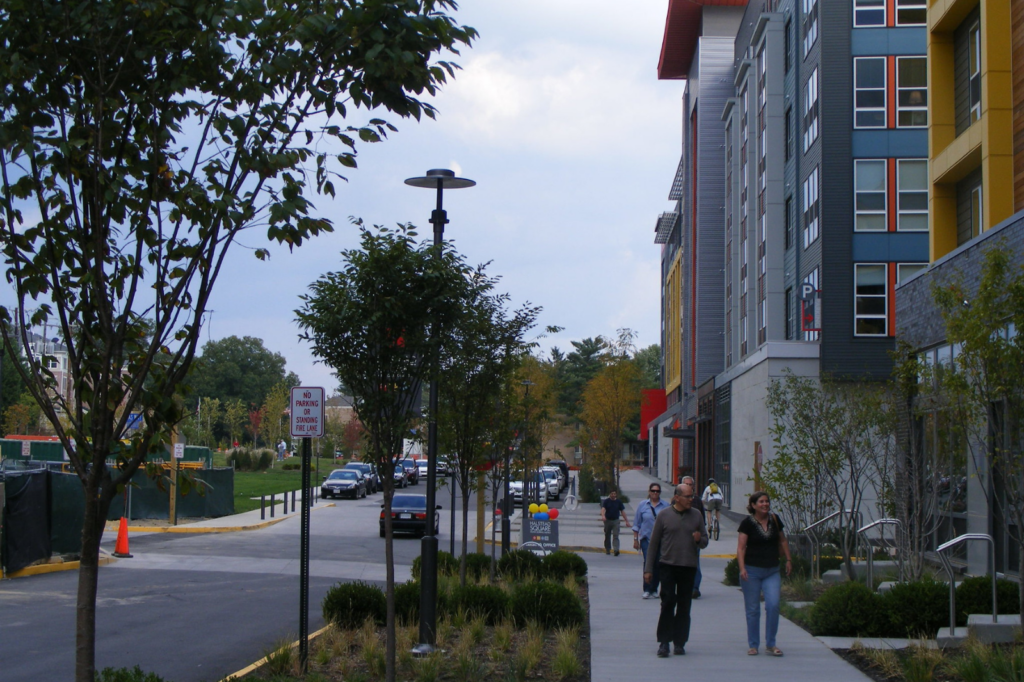 An assortment of people walk down a wide sidewalk near a brightly colored apartment building