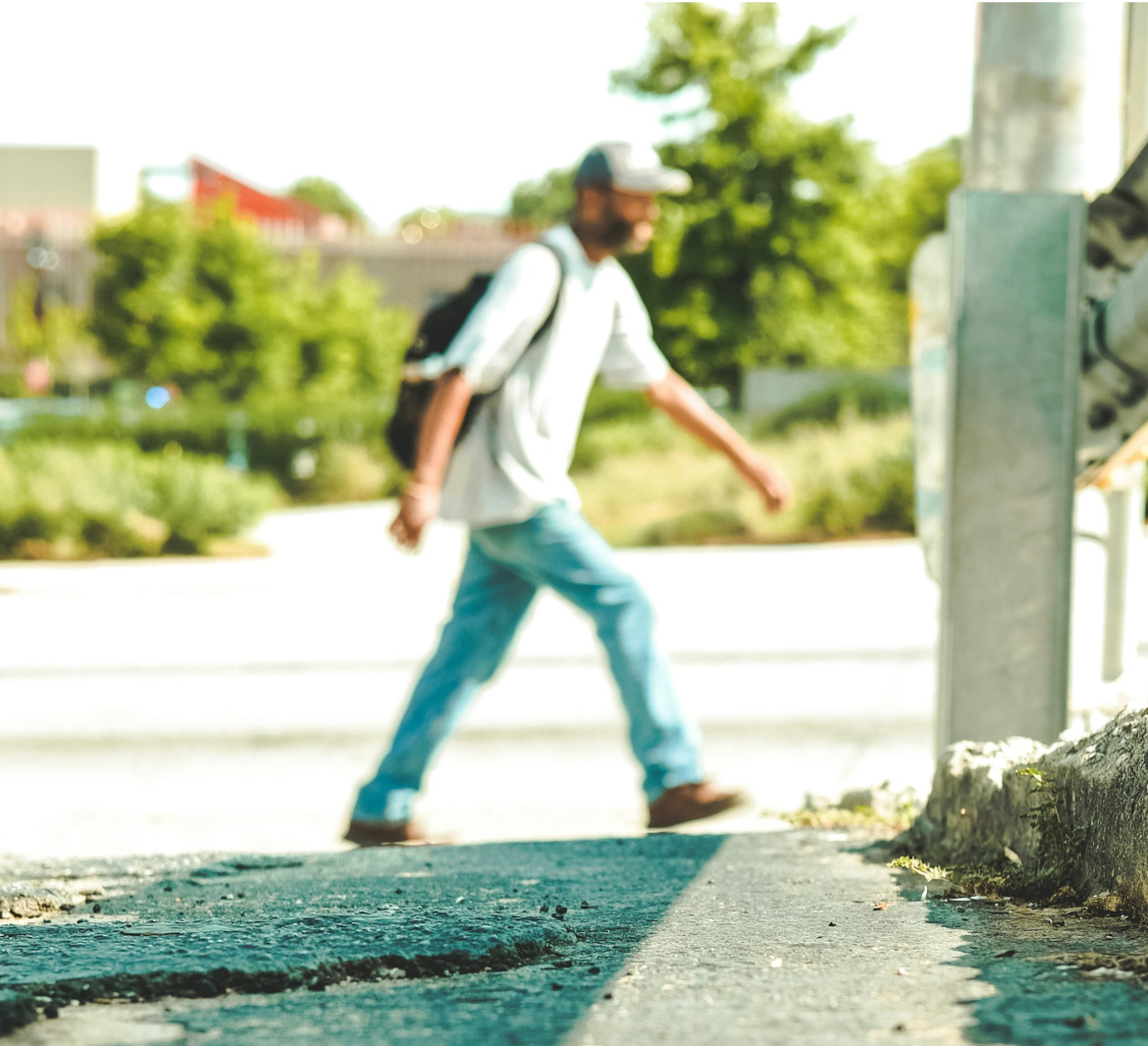 A man in jeans and a white t-shirt walks along the side of a wide, sunny street