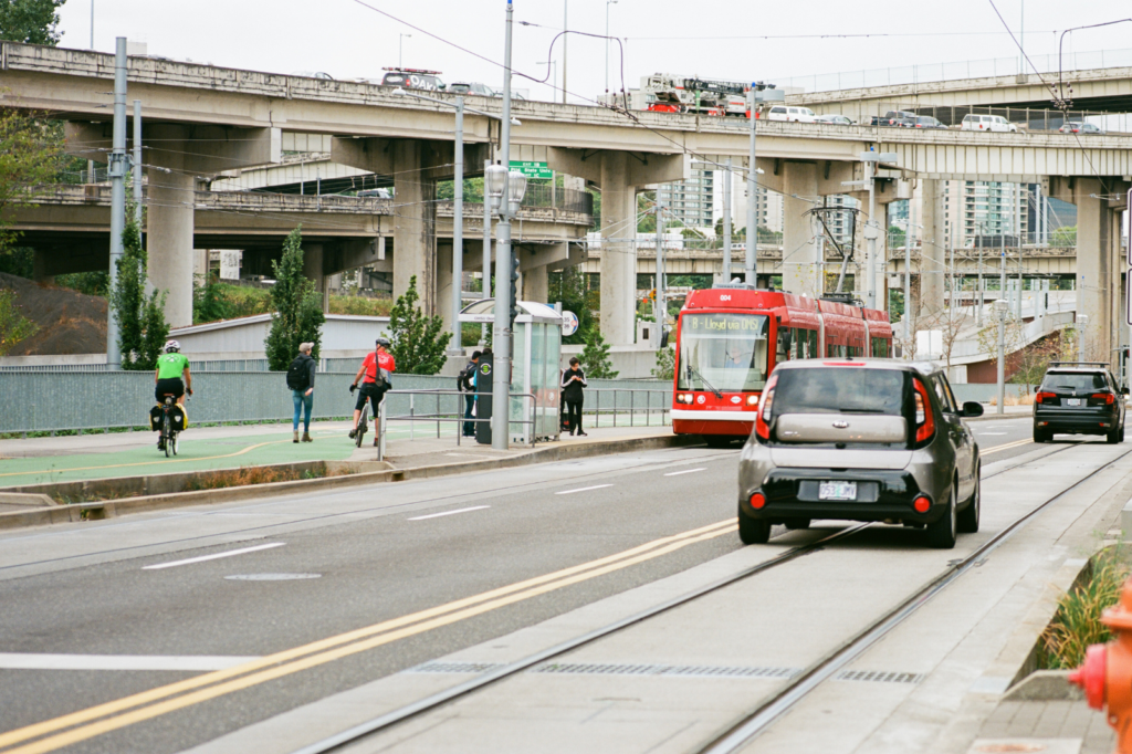 People cycle and walk down a green path near a transit stop.