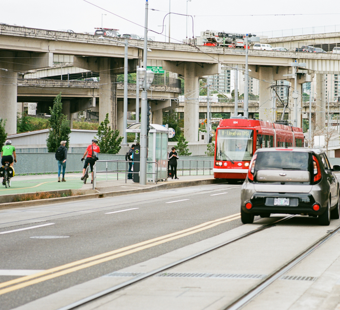 People cycle and walk down a green path near a transit stop.