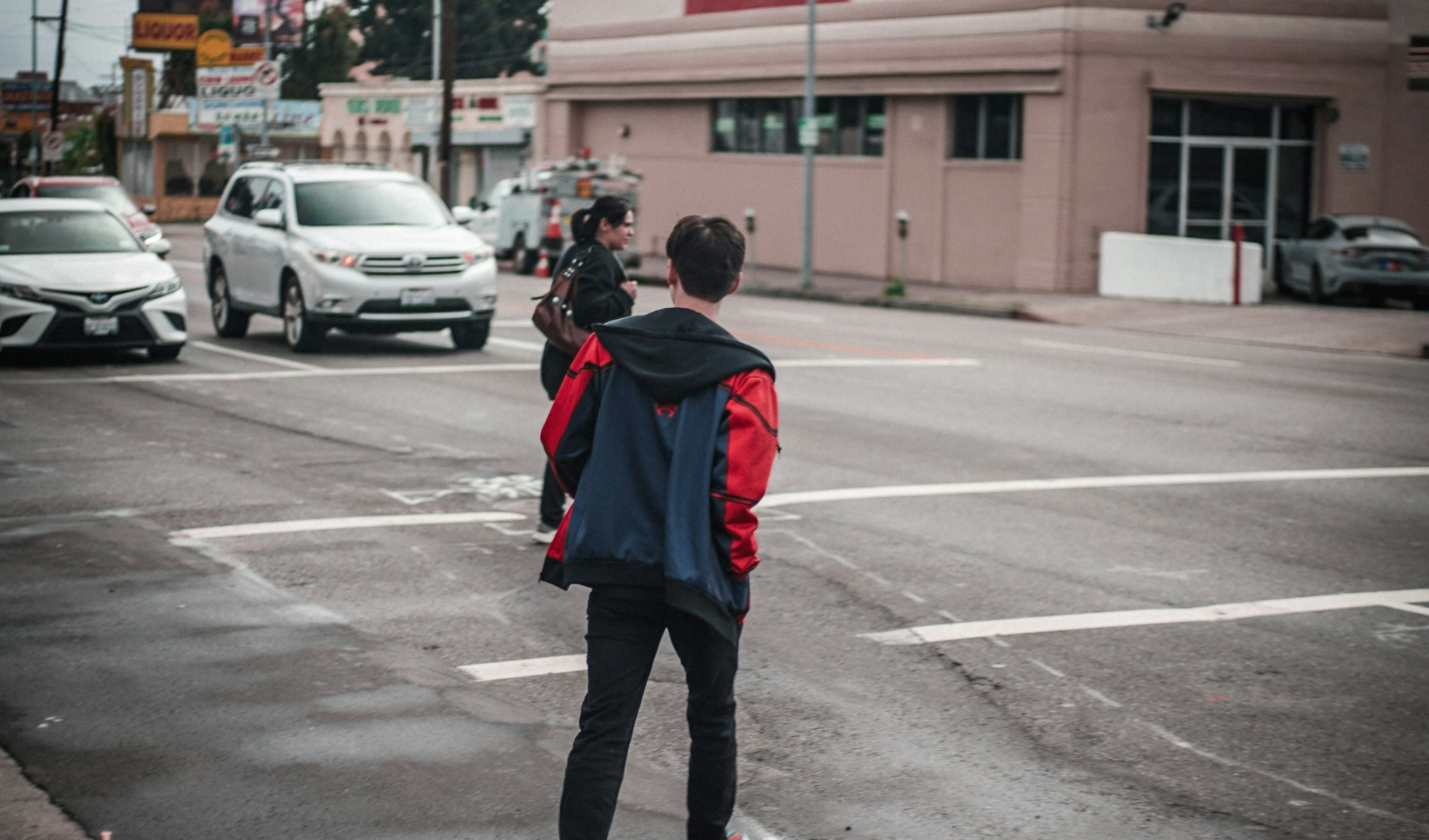 A young man and woman attempt to cross the street on a worn out crosswalk while two cars approach