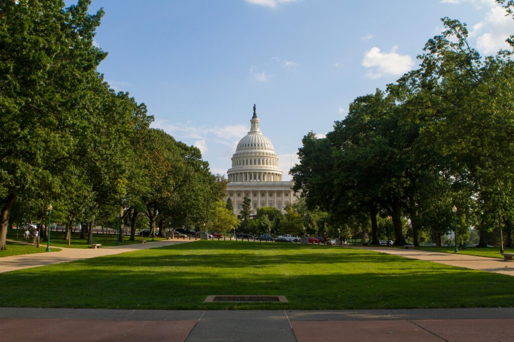 U.S. Capitol building in Washington, D.C., with the white dome centered in the background and green trees and lawn in the foreground.
