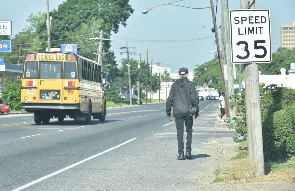 A man walks along the shoulder of a road with no sidewalk as a yellow bus passes. A 35 mph speed limit sign stands nearby.